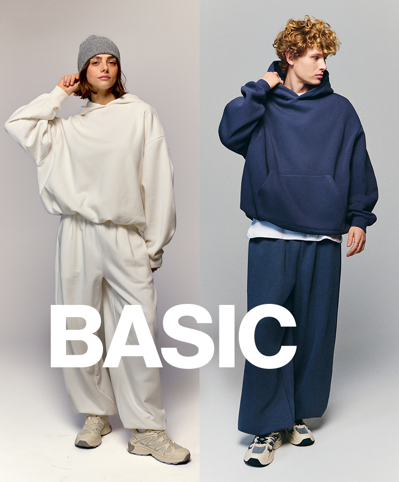 Two individuals wearing oversized, minimalist outfits are showcased side by side. One is dressed in a cream-colored hoodie and pants, paired with sneakers. The other wears a navy-blue hoodie and pants, also with sneakers. The word "BASIC" is prominently displayed in bold white text across the image.